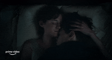 Movie gif. Harry Styles as Tom and Emma Corrin as Marion in My Policeman. He cuddles into her side as they lay in bed and she strokes his head lovingly.