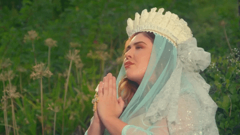 Music video gif. From video for Lizzo's Scuse Me, a woman wearing an ornate ruffled hat and veil presses her hands together in prayer with her eyes closed, while in a grassy field.