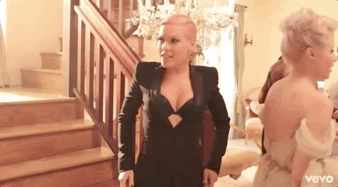 Celebrity gif. Wearing a black suit jacket with a black bra, Pink brings both hands up and fans her face, making a face as if to say, “It’s hot in here.”