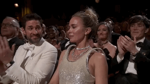 Oscars 2024 GIF. Emily Blunt, seated at the Oscars, shrinks back from the applause as if it were a bright light, then shaking off the shyness to confidently accept the praise.