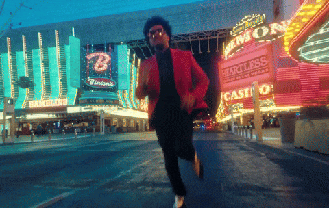 Celebrity gif. Singer The Weeknd (pronounced Weekend) runs wildly down the Las Vegas strip wearing a pink suit jacket and sunglasses. His arms flail wildly around him as he runs.