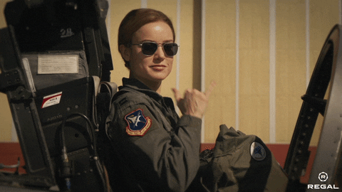 Disney gif. Brie Larson as Carol in Captain Marvel. She has aviators on and she's about to take off in a jet. She looks very cool as she flashes us the shaka and is ready for takeoff.
