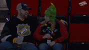 Video gif. Couple in the audience of a game look at each other giggling. The woman wearing a Christmas tree hat pretends to punch the man and he flinches, and laughs harder.