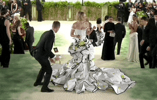Met Gala 2024 gif. Gigi Hadid holds one hand to her chest as six assistants swoop in from all around her to fix her dramatic mermaid-style skirt for camera. Hadid is wearing a cream colored full-length Thom Browne dress that is fitted through the drop waist that morphs into a full mermaid skirt with black edging, pale yellow roses, and green leaves that looks like a trench coat with lapels and a sleeve that hangs down from her hip.