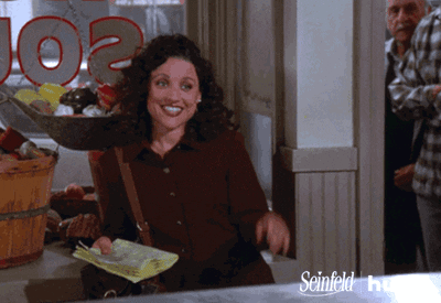 Seinfeld gif. Julia Louis Dreyfus as Elaine leans against a window and flutters her fingers waving hello, with a smile and raised eyebrows.