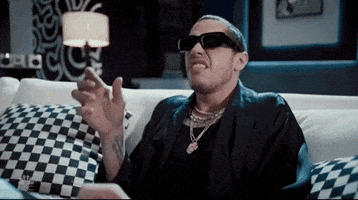SNL gif. Pete Davidson is wearing a robe and heavy jewelry and sunglasses as he sits indoors on a couch. He curls his nose up confused, and lifts his glasses to peer closer.