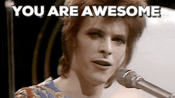 Celebrity gif. Young David Bowie stands at a silver microphone and points to us proudly saying, "You are awesome."