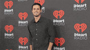 Celebrity gif. Ben Higgins is on the red carpet at the iHeartRadio Festival and he waves at us and smiles charmingly.