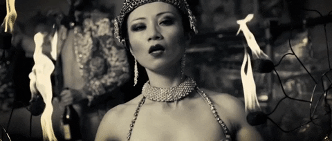Theater gif. Elegant Baroque Burlesque show woman with a blingy headpiece and a diamond choker and earrings looks at us seductively while wisps of flames dramatically surround her.