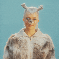 Video gif. Young woman wears a furry jacket and dog nose with platinum hair styled in tight coils and funky bangs against a teal background. She waves at us with both hands, smiling as she says, "Have a great day!'