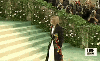 Met Gala 2024 gif. Anna Wintour poses for the cameras wearing a custom Loewe jacket adorned with colorful oversized flowers and a knowing smirk.