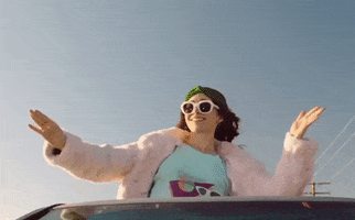 Movie gif. Cristin Milioti as Sarah in Palm Springs leaning out of a sunroof, smiling and dancing wearing sunglasses and a puffy jacket.