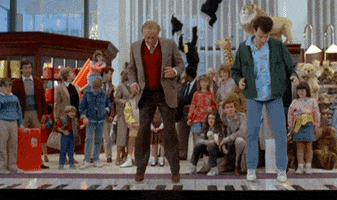 Movie gif. Tom Hanks and Robert Loggia as Josh and Mr. MacMillan in "Big" hopping on a light-up floor piano in FAO Schwarz as a crowd of parents and kids watch behind.