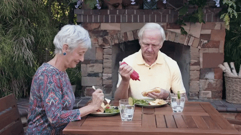 Video gif. Woman picks at her salad at a table as the man beside her shakes a bottle of ketchup, attempting to squeeze it onto a hot dog, but he misses and sprays ketchup all over his yellow shirt.