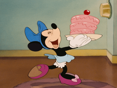 Cartoon gif. Minnie Mouse wears a light blue skirt, cornflower blue bow, and fuchsia shoes as she skips across a living space, bouncing up and down excitedly as she reaches her arms out, holding a pink birthday cake on a platter.