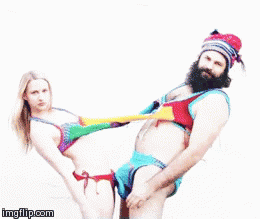 Video gif. A man and woman wear matching, rainbow, knit bikinis, which are attached to each other by extra pieces of wool fabric running between their chests and crotches. They rock back and forth and look at us. 