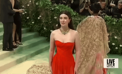 Met Gala 2024 gif. Closeup of Nell Diamond from above smiling for pictures wearing a cherry red Hill House Home sleeveless gown with a gold and diamond necklace, pink blush, and long straight hair with a structured bump at the top of her head.