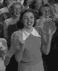 Video gif. Crowd of young women back in the 1950s clap widely and smile with excitement as they look up at someone in front of them.