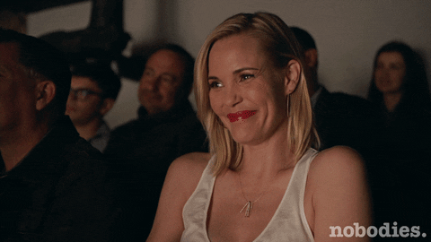 TV gif. Among an audience, on the show Nobodies, Leslie Bibb as Sam snickers quietly, raising her hand to cover her mouth.