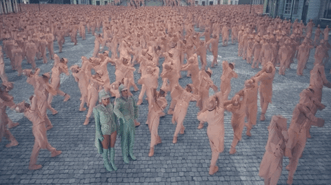 Music video gif. From video for "Me," Taylor Swift and Brendon Urie dressed in mint-colored uniforms, step in coordination with a mass of people dancing, dressed in pinkish-white uniforms.
