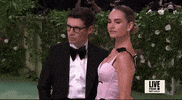 Met Gala 2024 gif. Closeup of Lily James posing for pictures. She dips a shoulder forward and back with a seductive expression. She's wearing a pale pink Erdem gown with a fitted bodice with a scalloped plunging neckline. A black vine with leaves detail weaves up and over her shoulders. 