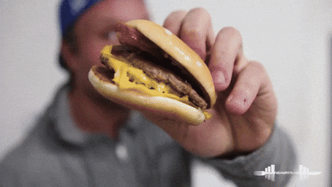 Video gif. Man holds a small simple burger up to us. He slowly moves the burger closer to his face, and then he looks down at it with excitement. He then chomps down onto the burger and stuffed it into his mouth.