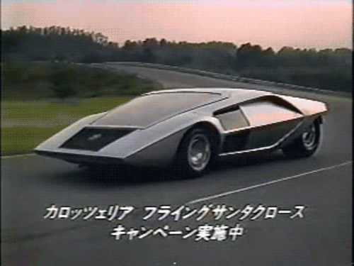 Video gif. An 80s futuristic concept car in Japan drives on the road at sunset. It is very angular and wedge-shaped and made of a silver metal. Japanese subtitles are on the screen. 