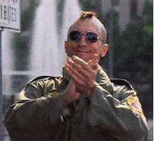 Movie gif. Robert De Niro as Travis Bickle in Taxi Driver, wearing aviator sunglasses and bulky jacket, smiles, and claps.