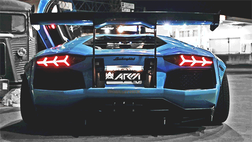 Video gif. From behind, we see a blue Lamborghini as sparks fly from its mufflers.