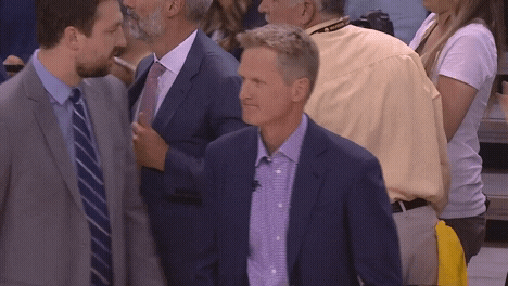 Sports gif. Steve Kerr, head coach of the Golden State Warriors, stands on the court looking around with a satisfied, closed lipped smile on his face. He stands still as others around him walk around quickly.