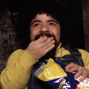 Video gif. A man is dazzled by something on TV and he has a bag of popcorn on his lap. He has a huge smile on his face as he shovels popcorn into his mouth. 