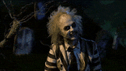 Movie gif. Michael Keaton as Beetlejuice sits in a graveyard while the lightning flashes. He holds his arms out and smiles as he says, “It’s showtime.” 