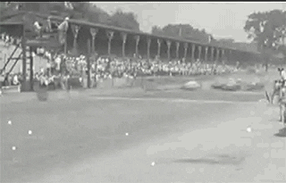 Video gif. Archival black and white footage of the Indy 500 shows vintage race cars speeding down a track as they kick up dirt.