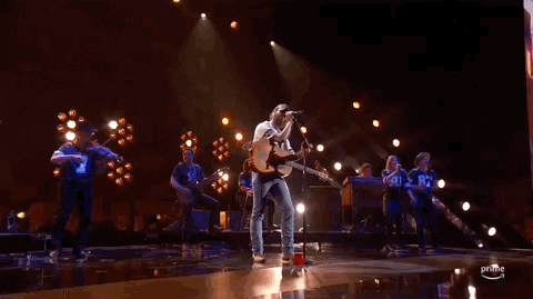 ACM Awards gif. Post Malone taps heel to the beat of the song with a guitar strapped around him as he sings into the standing mic while holding it with both hands and briefly shaking his head from left to right. 