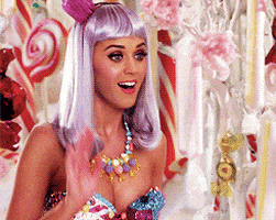 Celebrity gif. Standing in a room filled with candy, a smiling Katy Perry wearing a purple wig and a candy necklace waves and says, “Hi.”