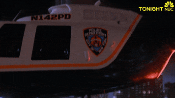 TV gif. A helicopter rises above the city and we see Andy Samberg as Jake in Brooklyn Nine-Nine come into screen holding onto the bottom of the chopper with one hand, looking calmly at us. Text, "Sup?"