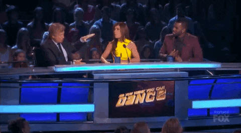 Reality TV gif. We see the judge panel on SYTYCD, where an older male judge gestures at Paula Abdul. Paula stands up and playfully starts pretending to punch him as he leans away from her.