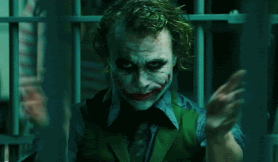 Move gif. Heath Ledger as the Joker in The Dark Knight smirks and glares forward at us while clapping his hands emphatically, standing in what appears to be a locked cell.