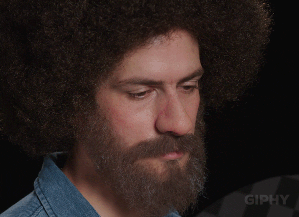 Video gif. Man dressed like Bob Ross has a sad look on his face as he looks down. He then slowly raises his head up and as he does, a smile spreads across his face and he looks around like he realizes everything is beautiful.
