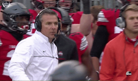 Sports gif. Urban Meyer walks down the sideline and falls facedown to the ground among other coaches and players watching the field with anticipation.