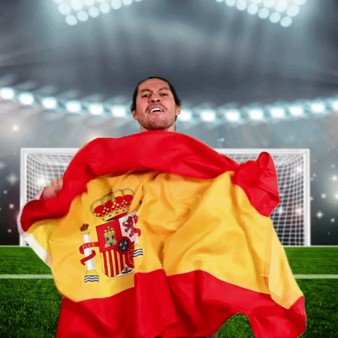 Digital compilation gif. Man wearing a red Spain National team jersey is edited to be standing on a soccer field with a goal and stadium lights behind him. He waves the Spanish flag up and down enthusiastically and screams towards us, "Vamos!'
