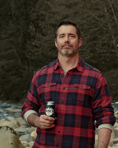 Ad gif. Man wearing flannel shirt, standing in front of a rocky creek, nods his head and raises his can of Busch beer toward us.