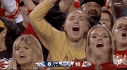 Video gif. Two women in the crowd at a Kansas City Chiefs game clap and scream excitedly in a crowd of fans, one making a number one symbol with her index finger and pumping her arm in the air as she screams and looks straight at us. 