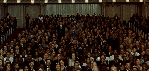 Video gif. Inside a ornate performance hall a full house rises in a standing ovation. 