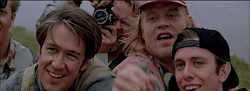 Movie gif. Philip Seymour Hoffman as Dusty in Twister leans forward between 2 other guys in a small crowd and gestures with his fingers as he says, "Food."