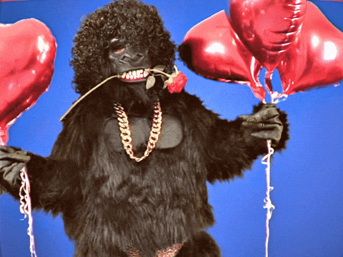 Video gif. Adult person in a full gorilla suit wearing a gold chain and skimpy underwear holds a few red heart-shaped balloons in each hand as they sway their hips and dance provocatively, a few stems of roses at their feet. 