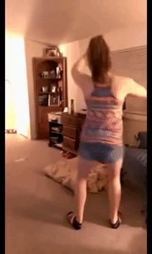 Video gif. A woman is facing away from us and is dancing mildly. All of a sudden, she bends and puts her hands on her knees to do an open-closed motion with her legs and hops away.