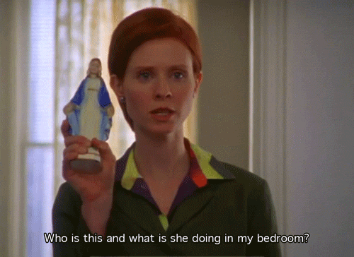 TV gif. Cynthia Nixon as Miranda Hobbes on Sex and The City has a very serious and irritated expression on her face as she holds up a small statue of Mother Mary. She says, “Who is this and what is she doing in my bedroom?”