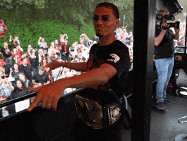 Sports gif. Amine Adli, a soccer player, is wearing a wrestling match belt and dances on stage with sunglasses on and a sassy expression.