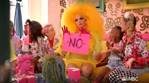 Reality TV gif. A person dressed in yellow with large yellow wig and pink gloves sits on a yellow couch with children dressed in extravagant outfits. The person holds up a pink sign with a large no written on it, drops it, then shrugs.